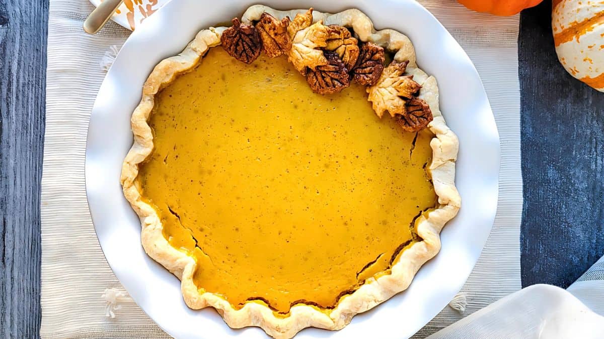 A pumpkin pie with a decorative crust sits on a white plate. The crust is wavy, and some parts feature leaf-shaped details. The pie is placed on a light-colored woven cloth, with a hint of autumn-themed decor nearby.