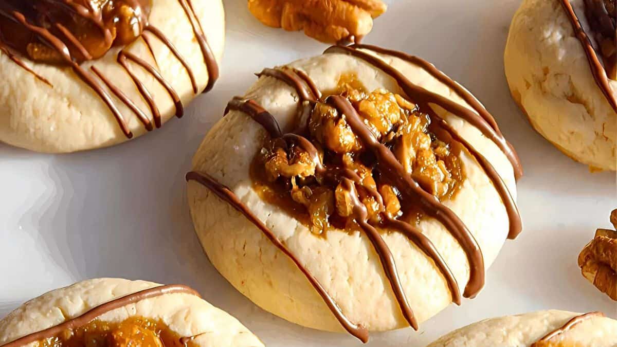 A close-up of pecan pie cookies drizzled with chocolate on a white plate. The cookies have a golden-brown, buttery base topped with a pecan and caramel mixture, surrounded by neat chocolate lines. Some pecan halves are visible in the background.