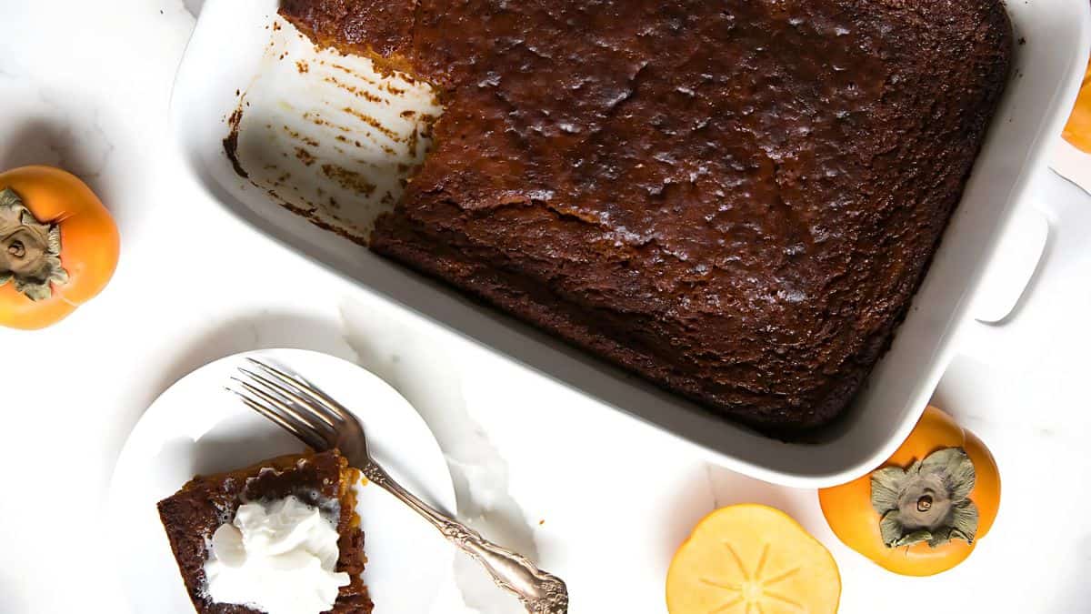 A sliced persimmon pudding in a white baking dish, with a single piece on a white plate garnished with whipped cream. Two whole persimmons and a sliced half are placed around the dish. A fork rests on the plate.