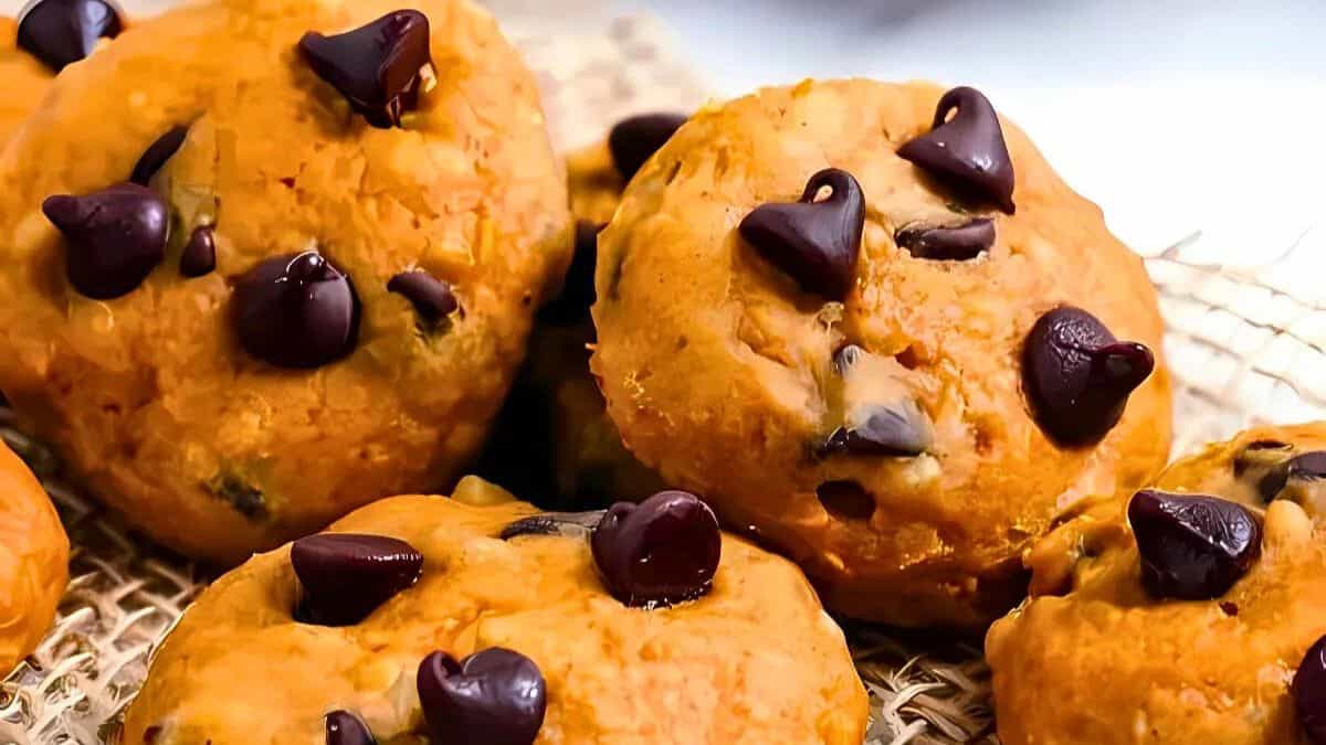 Close-up of a pile of cookies with a golden-brown texture, topped with scattered dark chocolate chips. The cookies are placed on a beige woven surface, highlighting their homemade appearance.