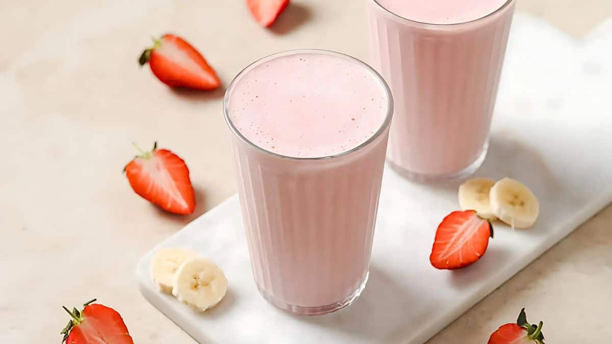 Two glasses of pink smoothie on a white surface, surrounded by slices of fresh strawberries and bananas. The light background enhances the vibrant colors of the fruits, creating a refreshing and inviting scene.