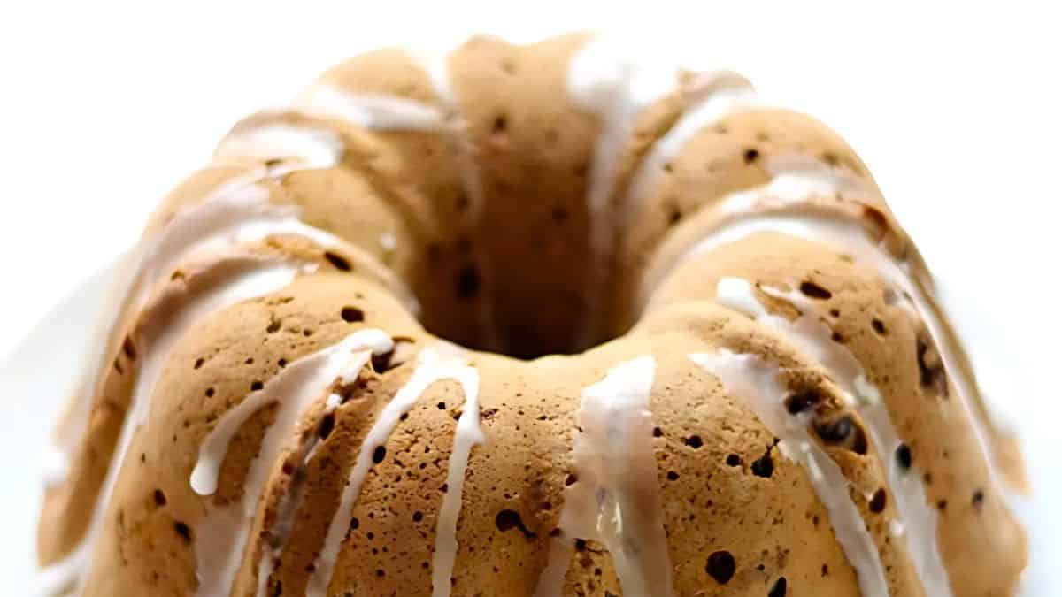 A close-up of a glazed bundt cake with a light brown color, featuring small chocolate chips. The cake is drizzled with white icing, set against a plain white background.