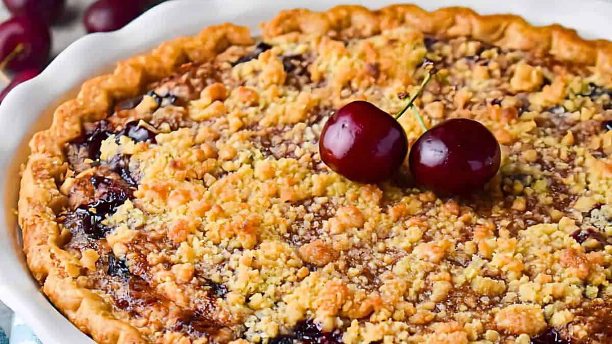 A freshly baked cherry crumble pie in a white dish, topped with two whole cherries. The pie features a golden-brown crust and a textured, crumbly topping with visible cherry filling.