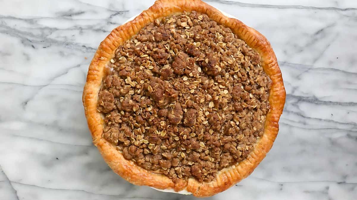 A freshly baked apple pie sits atop a marble surface. The pie has a golden, flaky crust and is topped with a generous oatmeal crumb streusel.