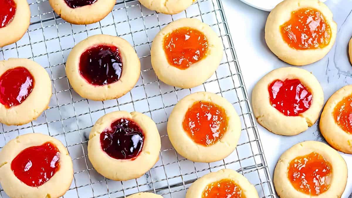 A cooling rack filled with thumbprint cookies topped with various fruit jams, including strawberry, blueberry, and apricot. The cookies are arranged neatly on a marble countertop, showcasing their vibrant jam centers.