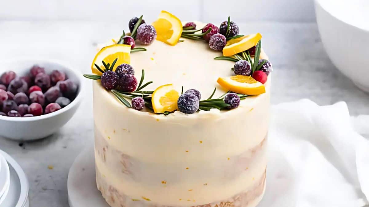A frosted cake topped with sugared cranberries, orange slices, and rosemary sprigs, placed on a white platter. In the background, there's a bowl of cranberries and a white cloth on a marble surface.
