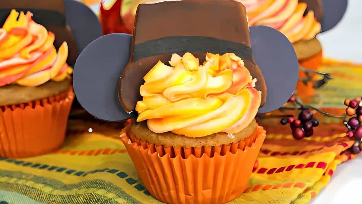 Cupcakes with orange and yellow swirled frosting, topped with chocolate hats resembling Mickey ears, are placed on a colorful striped cloth with small decorative berries.