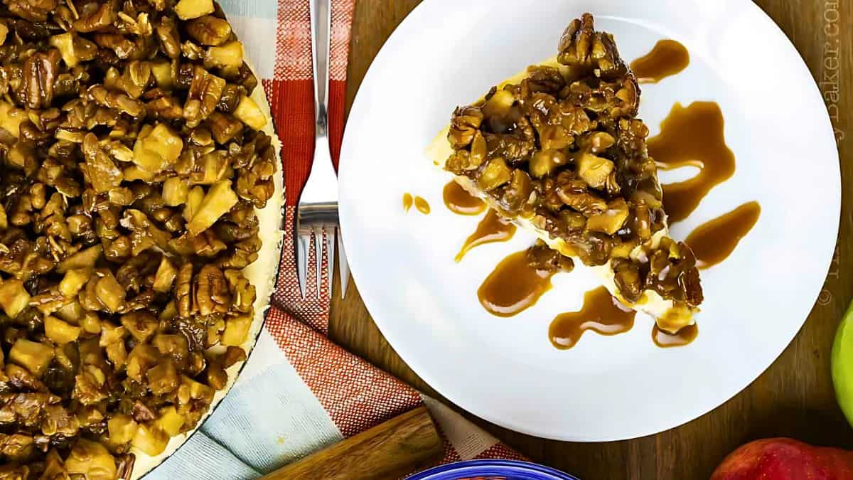 A slice of caramel pecan cheesecake on a white plate, drizzled with caramel sauce. The whole cheesecake, topped generously with nuts and caramel, is partially visible. A blue bowl and a red apple are nearby on a wooden table.