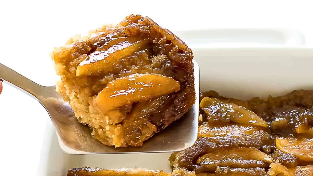 A close-up of a serving of caramelized pineapple upside-down cake being lifted from a baking dish with a metal spatula. The cake has a golden-brown top with slices of pineapple visible.