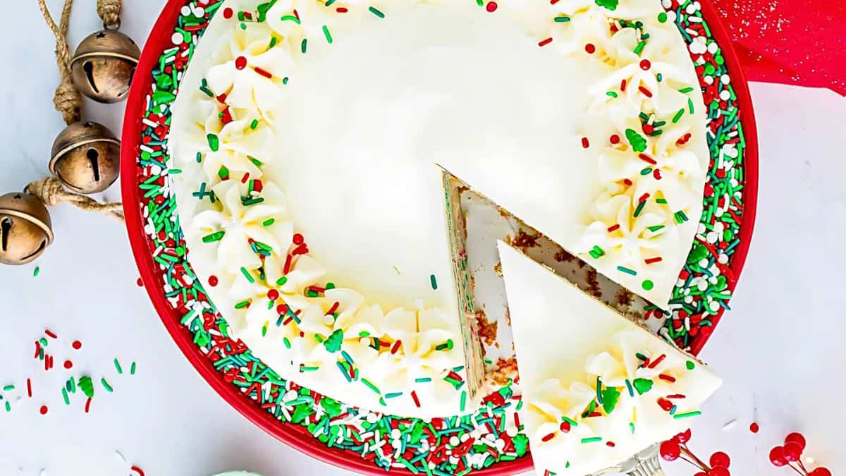 A festive round cake with white frosting, topped with red and green sprinkles. One slice is cut and being lifted, revealing the moist interior. The cake is on a red plate, and there are decorative bells nearby on a white surface.