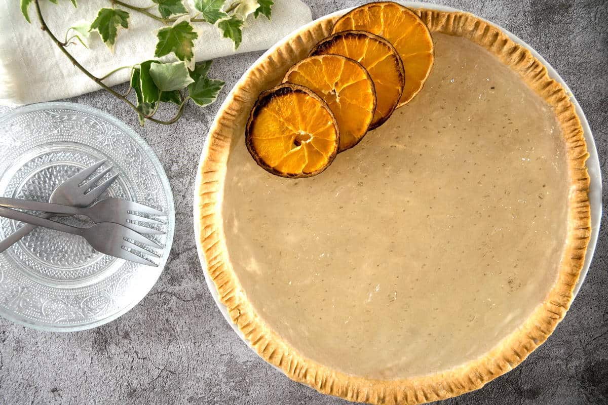 A round pie with a smooth, creamy beige filling is garnished with four dried orange slices on one side. Next to the pie are two forks resting on a decorative glass plate, and a white napkin with green leaves is on the side.
