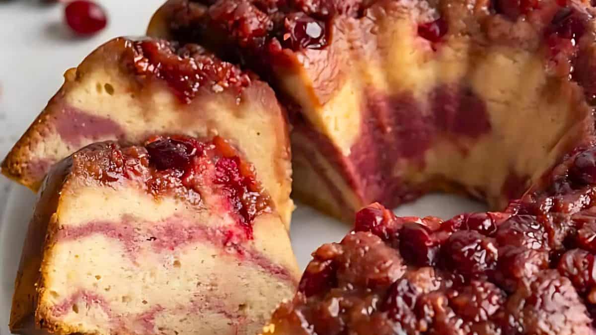 A close-up of a cranberry coffee cake with a rich, marbled texture. The cake is topped with a glaze and whole cranberries, and a slice is cut to show its interior layers.