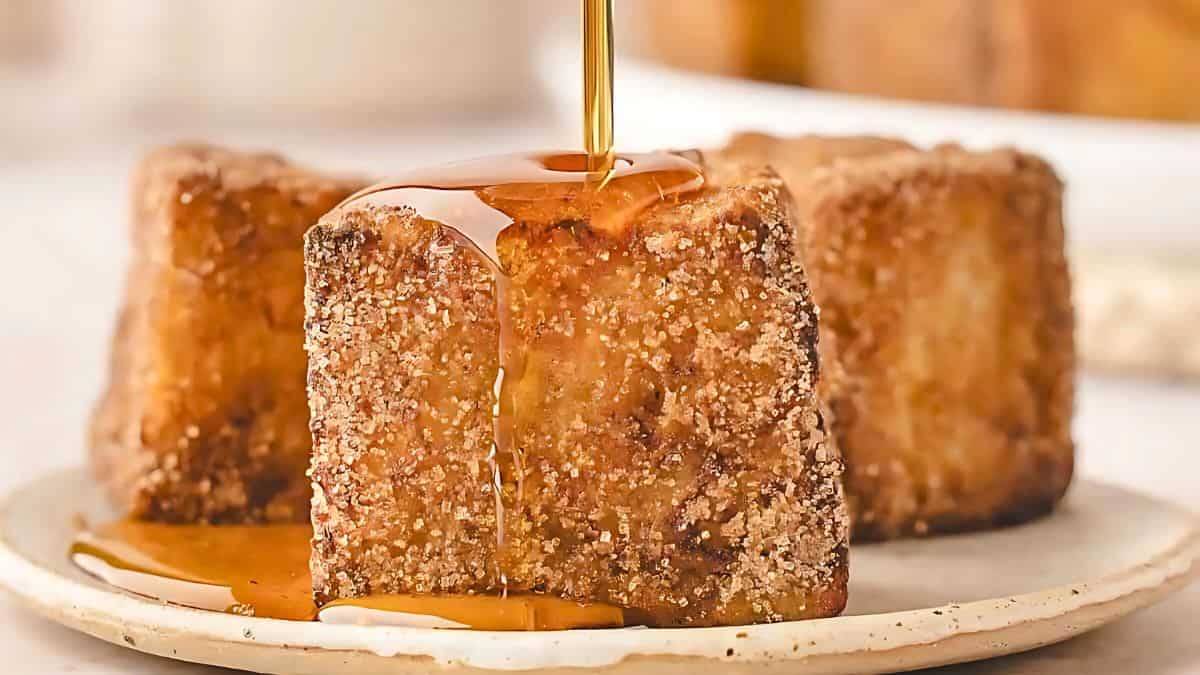 A close-up of a cinnamon sugar-coated French toast cube on a plate, being drizzled with syrup. The background is softly blurred, highlighting the crispy texture and golden-brown color of the cube.