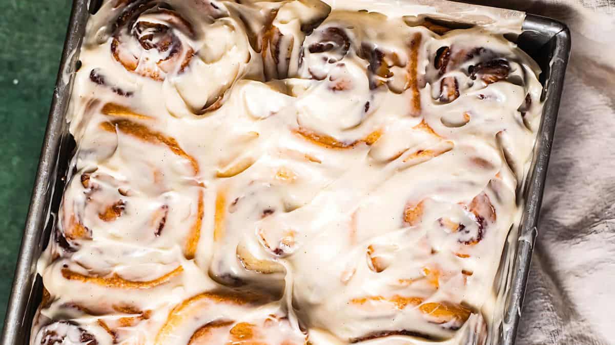 A tray of freshly baked cinnamon rolls covered in creamy white icing, with the rolls slightly visible beneath the glaze. The tray is on a green surface with a hint of a white cloth on the side.