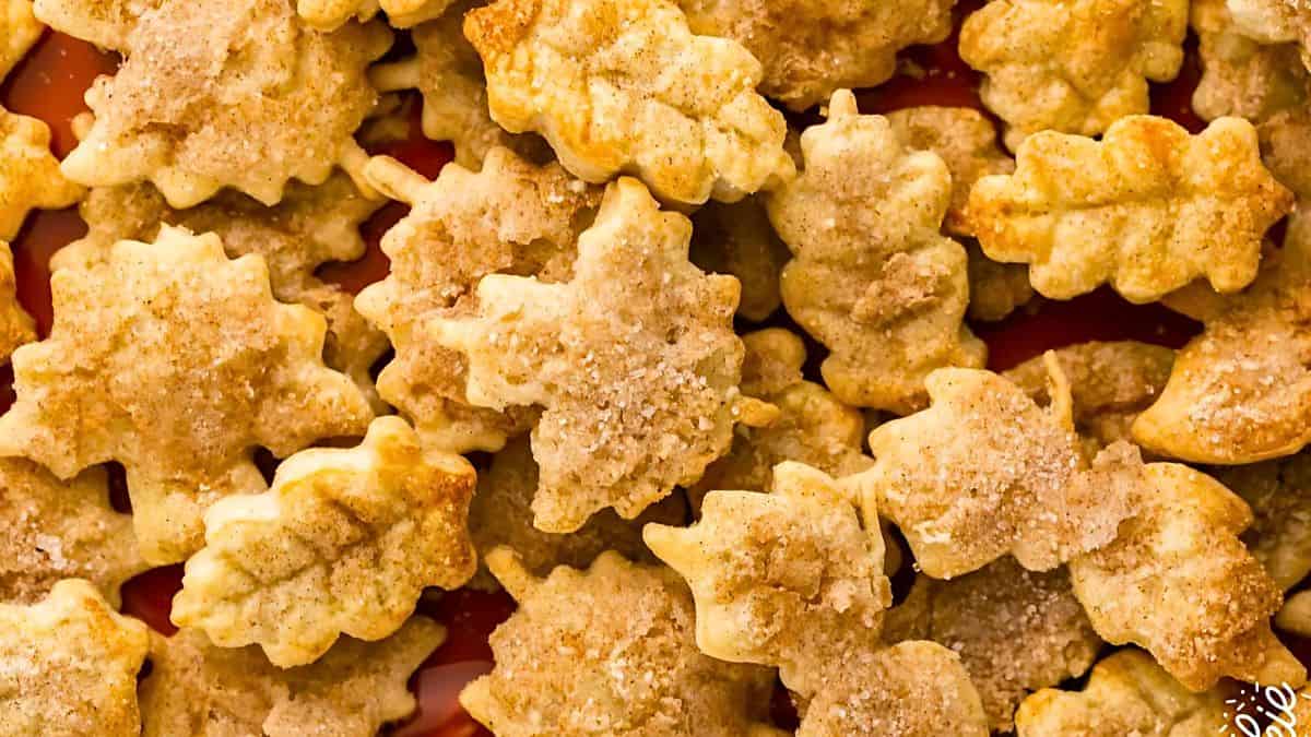 A close-up of leaf-shaped cookies topped with sugar and cinnamon. The cookies are in various shades of golden brown, highlighting the textured leaf patterns against a red background.