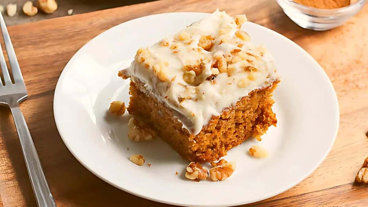 A slice of carrot cake with creamy icing and walnut pieces on top, served on a white plate. A fork rests nearby on a wooden table, with a small bowl of additional walnuts in the background.