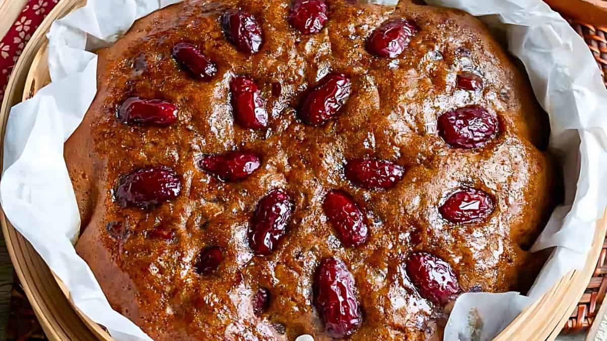 A round brown cake topped with glossy red dates, resting on white parchment paper. The cake has a shiny, moist texture and is presented on a wooden board.