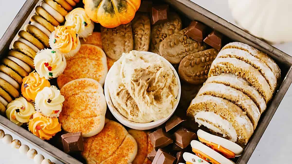 A dessert tray featuring a variety of cookies, cake slices, and chocolates, with a bowl of creamy dip in the center. Decorations include small pumpkins, adding a festive, autumnal touch.