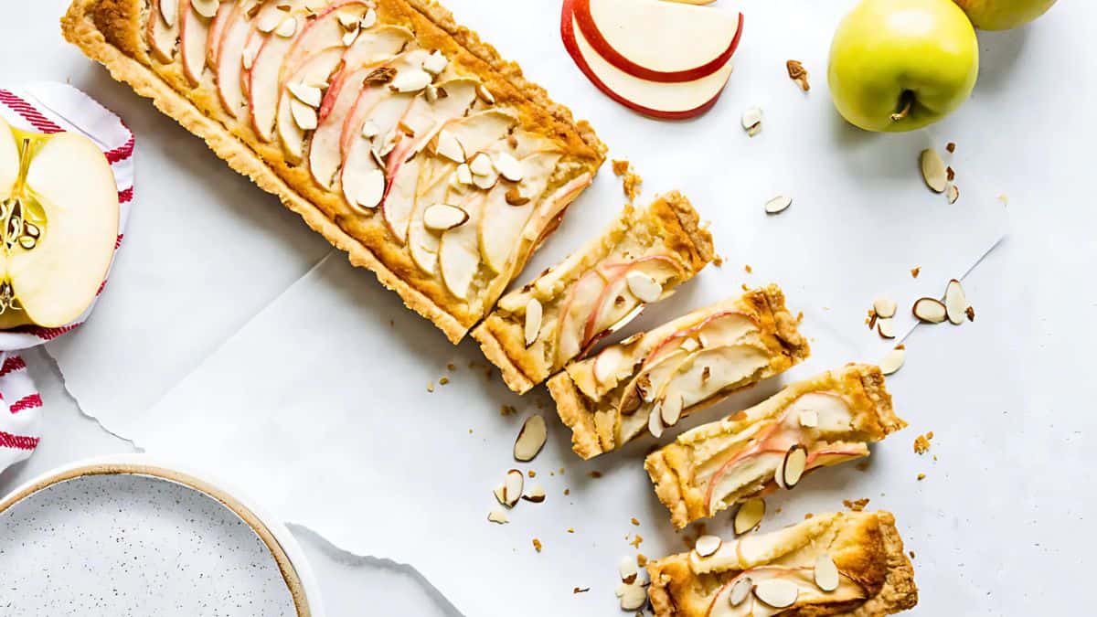 A rectangular apple tart with sliced almonds is cut into pieces on a white surface. Fresh green apples and red apple slices are scattered around. A striped cloth is partially visible on the side.