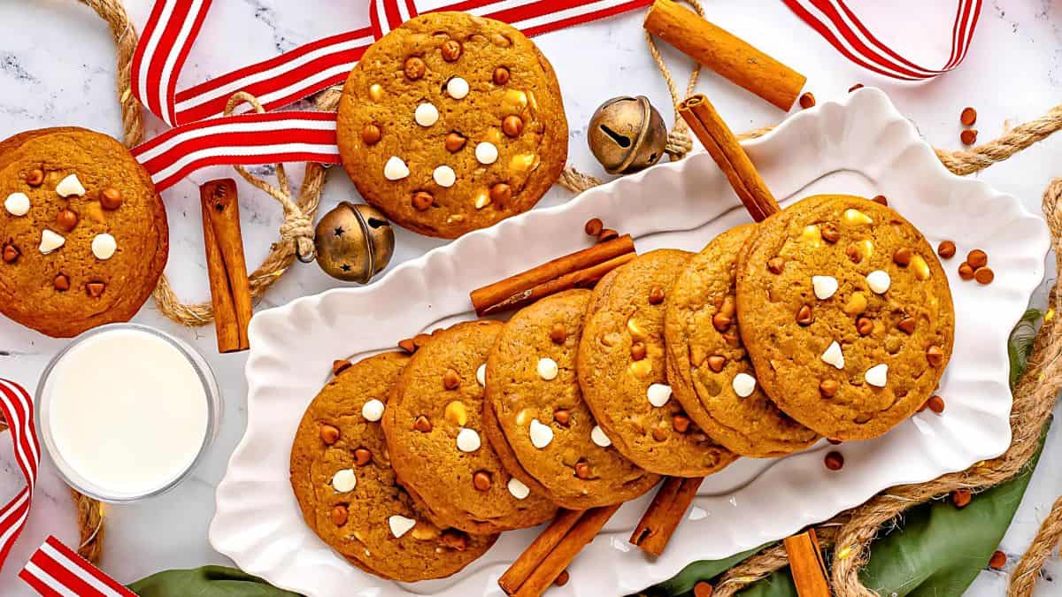 A plate of pumpkin cookies with white chocolate chips and cinnamon chips, surrounded by cinnamon sticks, red and white ribbons, and jingle bells. A glass of milk is placed nearby on a marble surface.