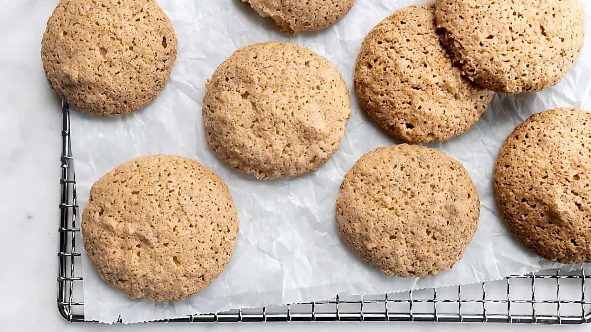 Seven almond cookies on parchment paper placed on a wire cooling rack. The cookies are round, golden-brown, and have a slightly rough texture. They sit on a light, marble surface.