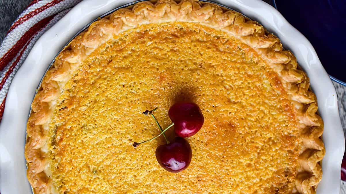 A golden-brown pie with a decorative crust sits in a white pie dish. Two red cherries are placed in the center of the pie. A striped cloth and a blue cup are partially visible in the background.