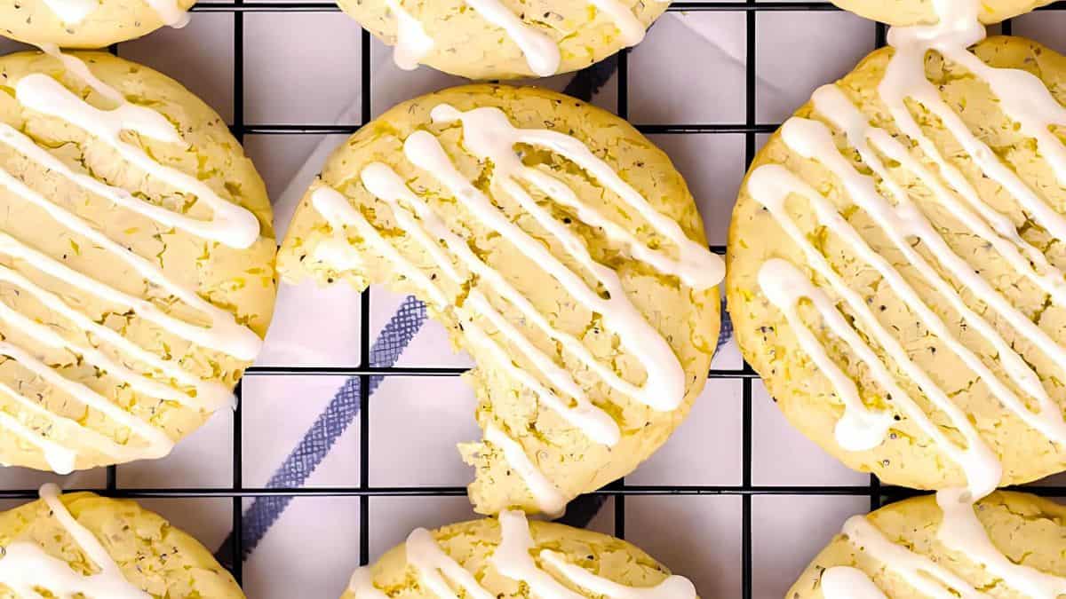 Lemon cookies with white drizzle icing are arranged on a cooling rack. One cookie has a bite taken out of it, showcasing its soft texture. The cookies are light yellow, and the icing is artfully drizzled.