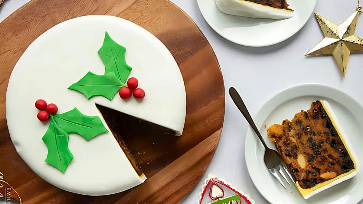 A round fruitcake with white icing is decorated with green holly leaves and red berries. A slice is placed on a plate next to a fork. A gold star ornament and a small gift box are nearby on a white surface.