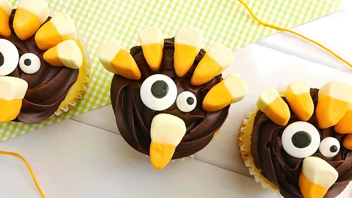 Cupcakes decorated with candy corn and candy eyes to resemble turkeys. The cupcakes have chocolate frosting and are arranged on a green and white checkered cloth. Yellow strings are placed around them.