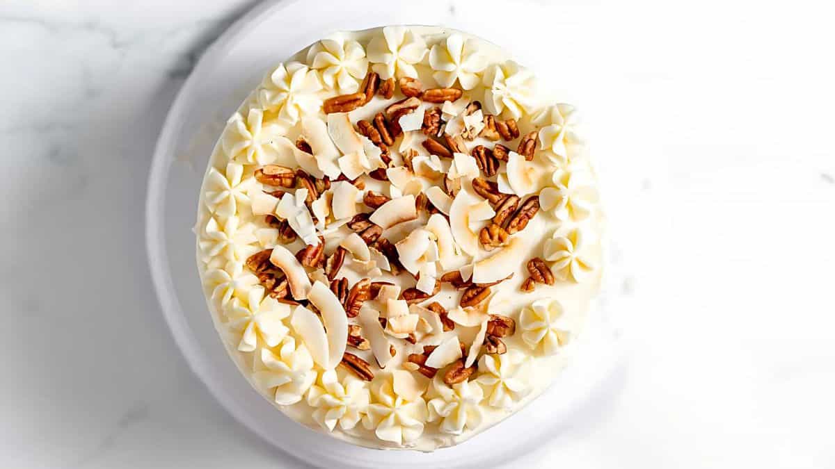 A top view of a cake with creamy frosting, decorated with chopped pecans and large coconut flakes. The frosting forms a ring of decorative swirls around the edge, creating an elegant presentation against a white background.