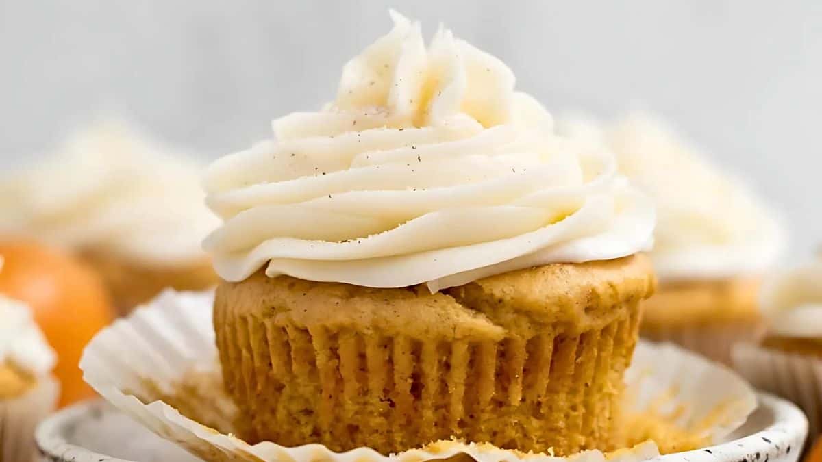 A close-up of a cupcake with creamy white frosting swirled on top, set in a crinkled paper liner. The cupcake has a light brown base, suggesting a warm flavor like vanilla or spice. The background is softly blurred with hints of other cupcakes.