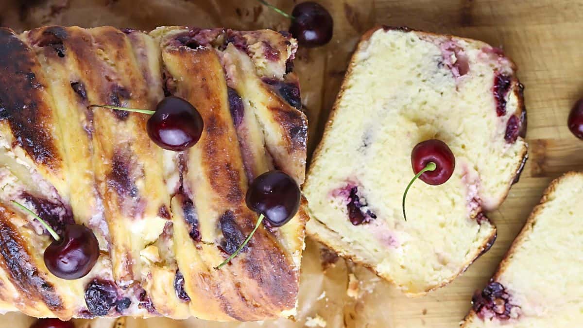 Freshly baked cherry bread with a golden crust is topped with whole cherries. Slices reveal a soft, moist interior dotted with cherry pieces. The bread is on a wooden surface, with a few additional cherries scattered around.