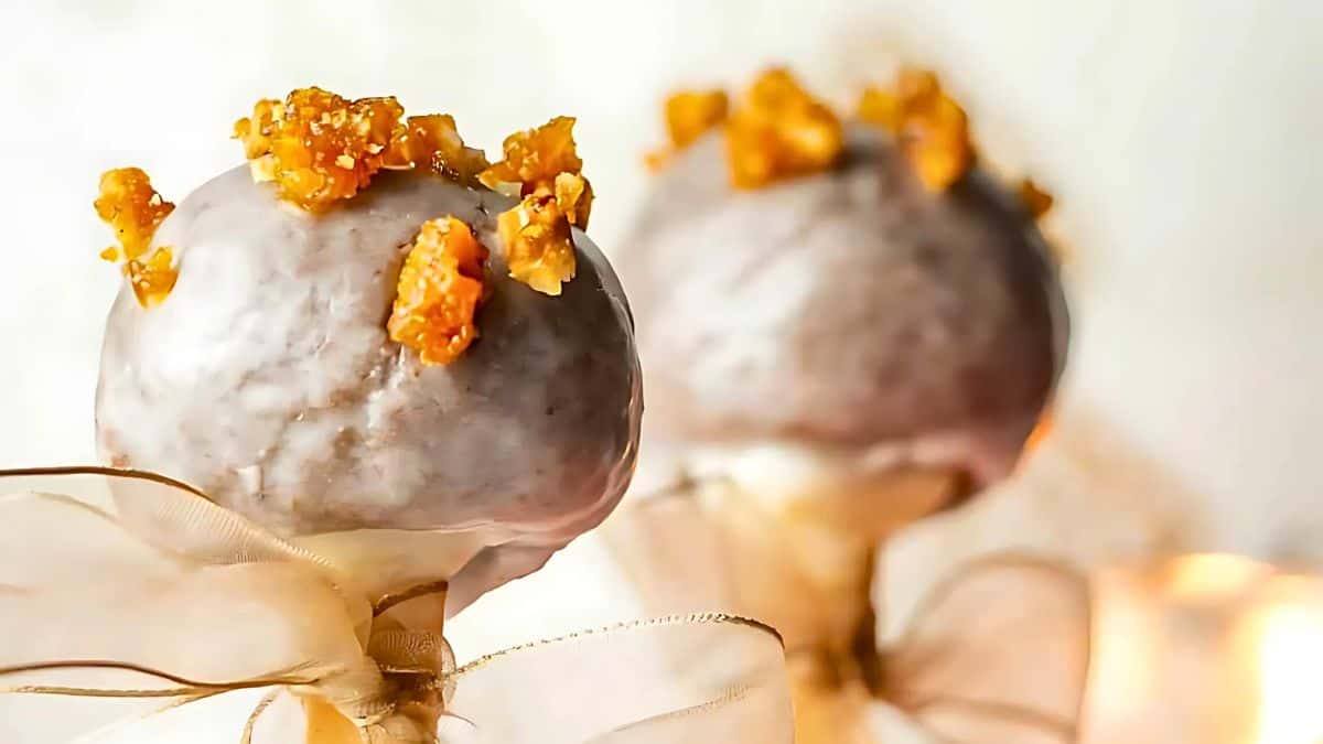 Two cake pops covered in white icing, topped with small pieces of caramelized nuts, are adorned with beige ribbon bows, set against a blurred light background.