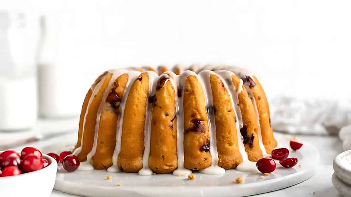 A bundt cake drizzled with white icing sits on a white plate. The cake is dotted with cranberries, and a small bowl of cranberries is nearby. Two bottles of milk are blurred in the background.