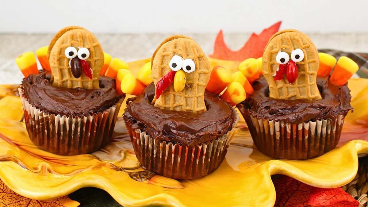Three chocolate-frosted cupcakes are decorated to resemble turkeys, each with a cookie for a head, candy eyes, a candy corn beak, and colorful candy feathers. They sit on a decorative autumn leaf platter.