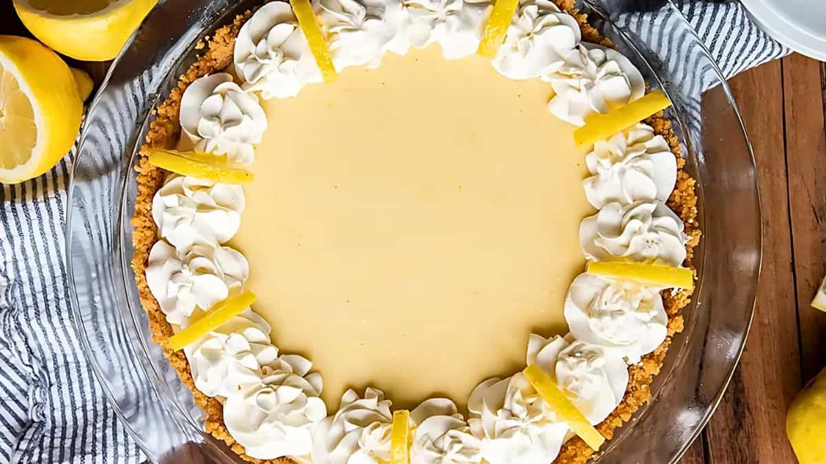 A lemon pie with a creamy filling in a crumbly crust sits on a wooden table. It's topped with swirls of whipped cream and garnished with lemon wedges. Fresh lemons and a striped cloth surround the pie.