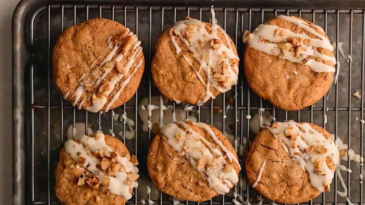 Six freshly baked cookies are placed on a cooling rack. They are topped with a drizzle of white icing and sprinkled with chopped nuts, which adds texture and contrast to the golden-brown cookies. The rack is set on a metal baking sheet.