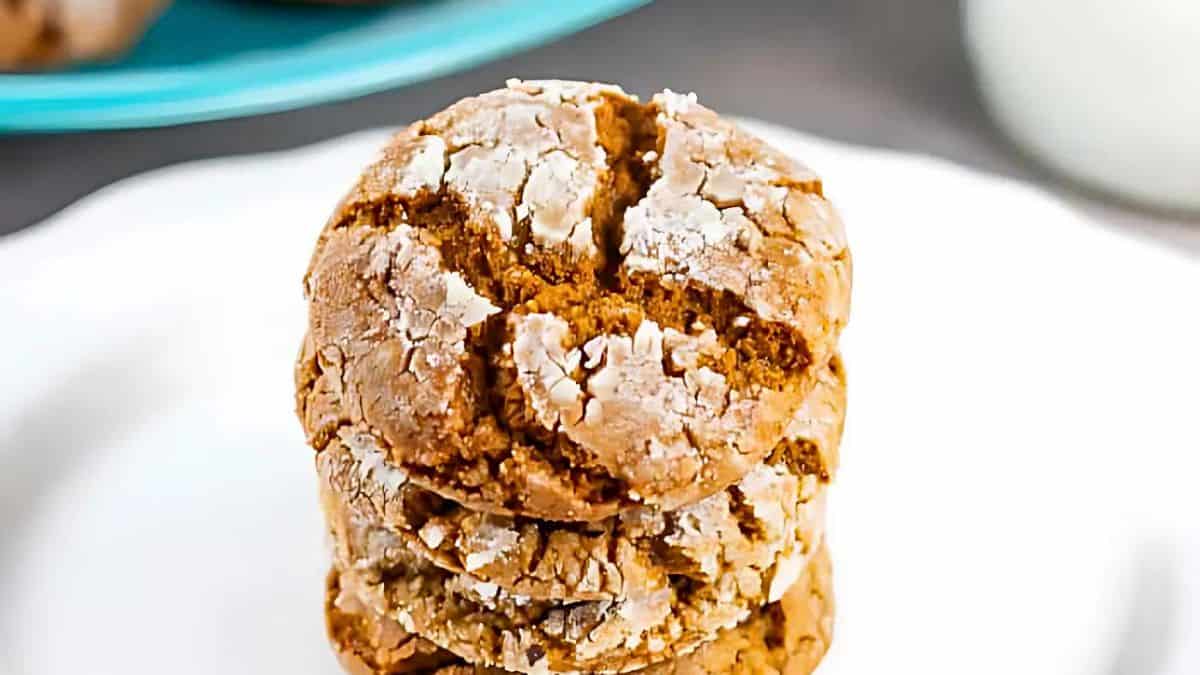 A stack of three cracked cookies dusted with powdered sugar sits on a white plate. The background includes a blurred hint of a blue dish and a glass of milk.