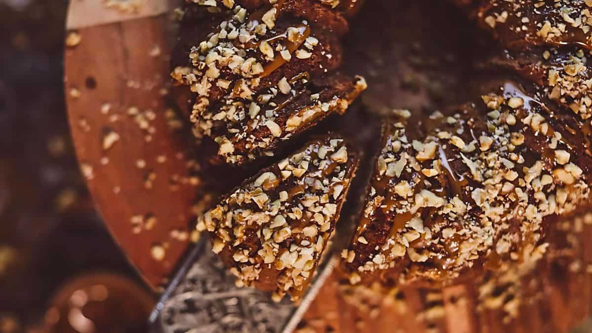 A close-up of a round cake topped with chopped nuts. A knife is partially inserted, cutting a slice from the cake, which is displayed on a wooden surface. The rich texture and warm tones suggest a caramel or chocolate drizzle.