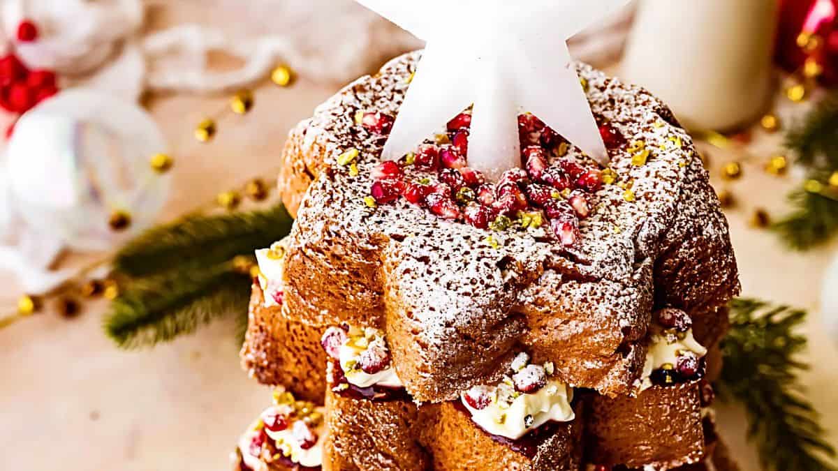 A festive star-shaped cake decorated with powdered sugar, pomegranate seeds, and cream, topped with a white star ornament. Greenery and golden beads surround it, creating a holiday ambiance.