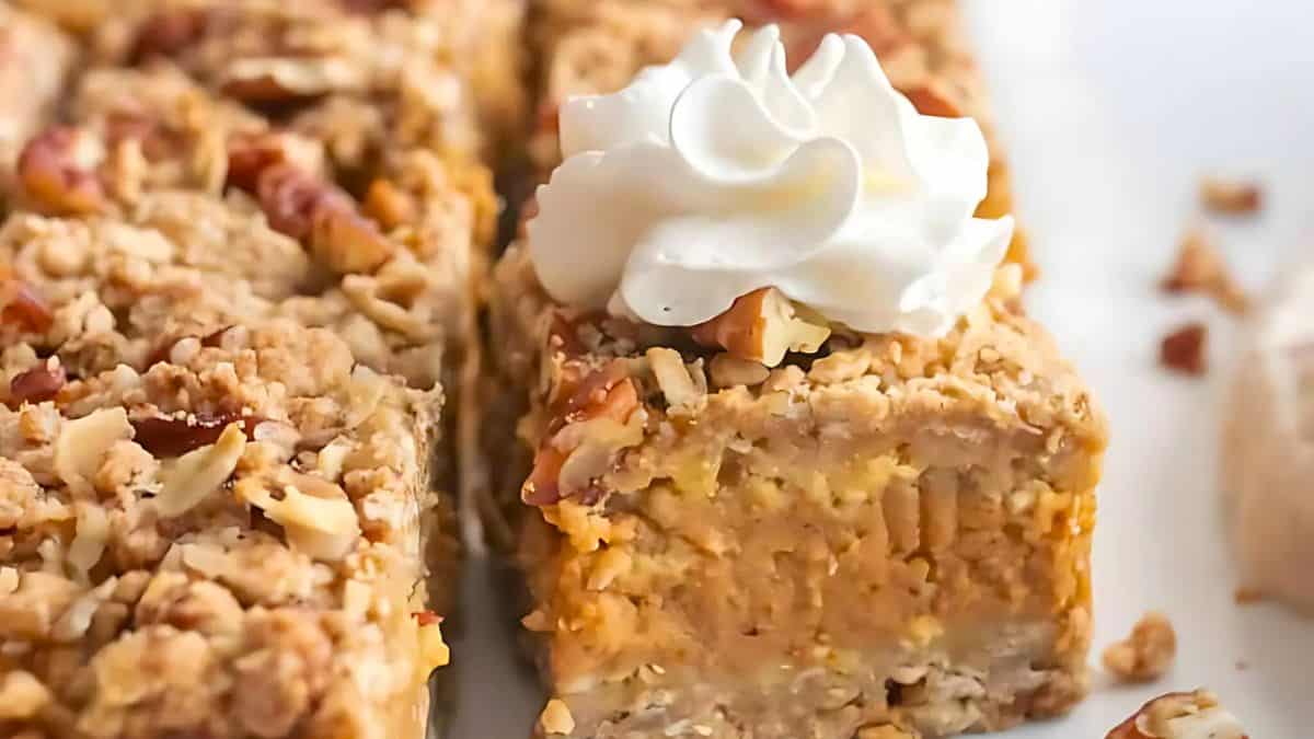 A close-up of sliced pumpkin cheesecake bars topped with a crumbly pecan crust and a swirl of whipped cream on top. The bars have a creamy texture and are garnished with chopped nuts.