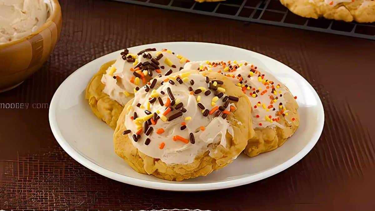 A plate of three cookies topped with white frosting and sprinkled with chocolate and orange sprinkles, resting on a brown table. In the background, a cooling rack with more cookies and a bowl are partially visible.
