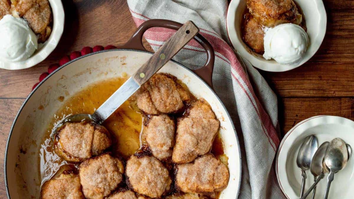 A baking dish filled with golden brown apple dumplings covered in syrup, with a serving spoon. Two bowls with apple dumplings topped with vanilla ice cream are beside it. A striped cloth and silver spoons are on the wooden table.