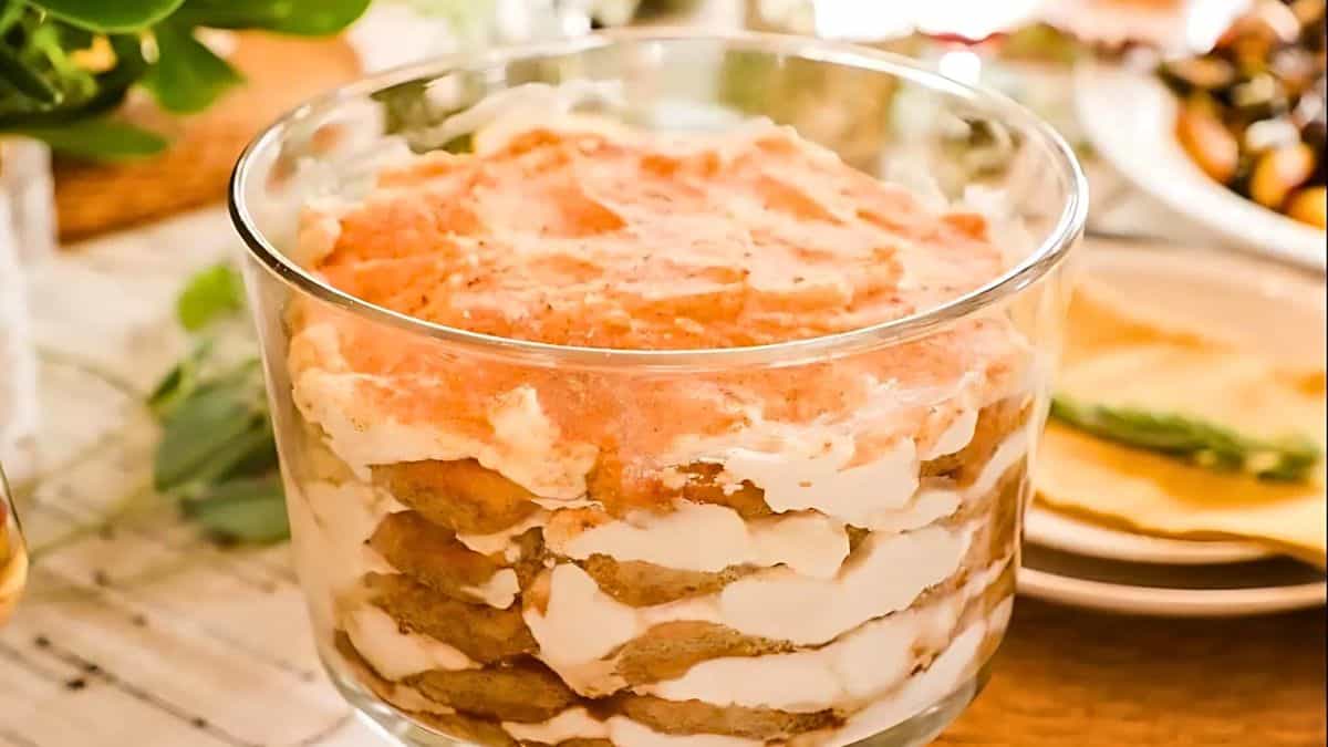 A glass dish filled with layers of creamy dessert topped with a light pink sauce. The dessert is set on a wooden table with other dishes partially visible in the background.