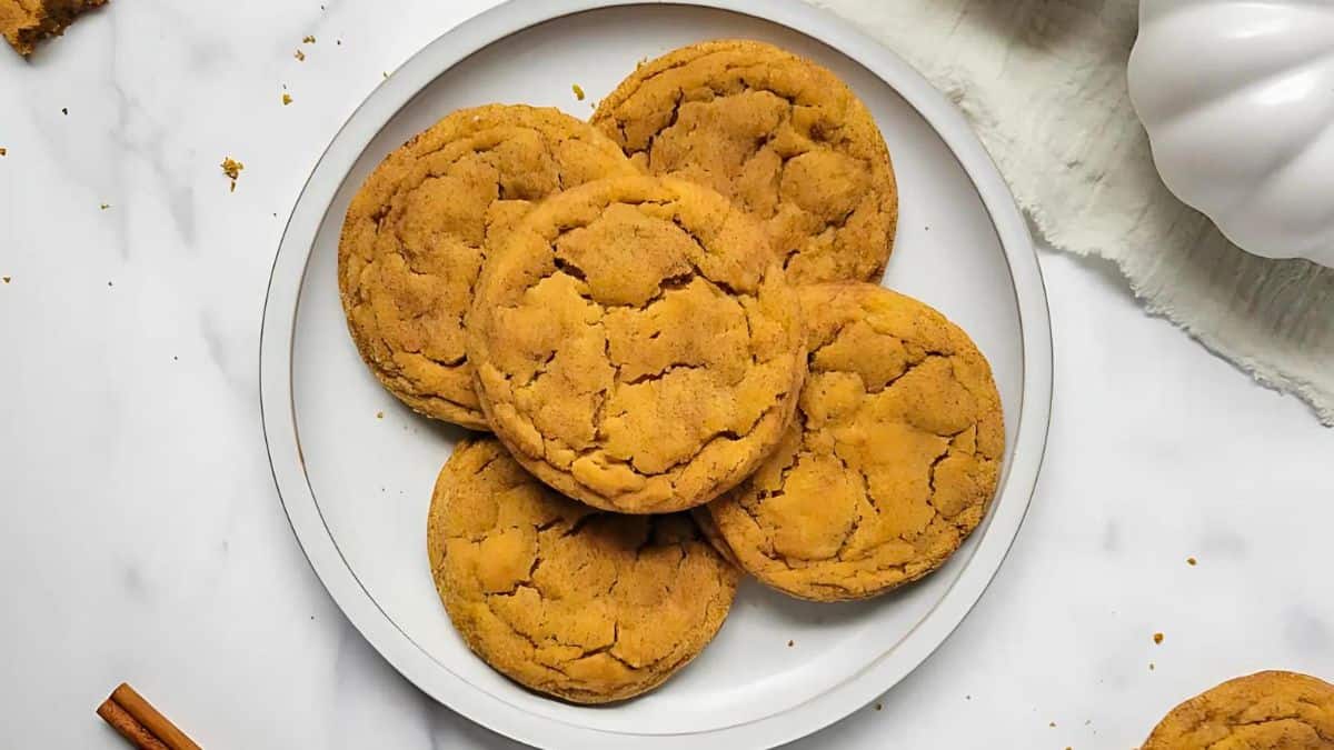 A plate of five pumpkin cookies is arranged in a circle on a white surface. The cookies are golden-brown with a cracked texture. A white pumpkin decor and a piece of cinnamon stick are partially visible nearby.
