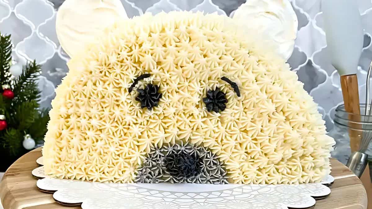 A bear-shaped cake covered in textured vanilla icing, with black and gray icing for the eyes, nose, and mouth. The cake sits on a decorative white paper on a wooden surface. A small Christmas tree and kitchen utensils are in the background.