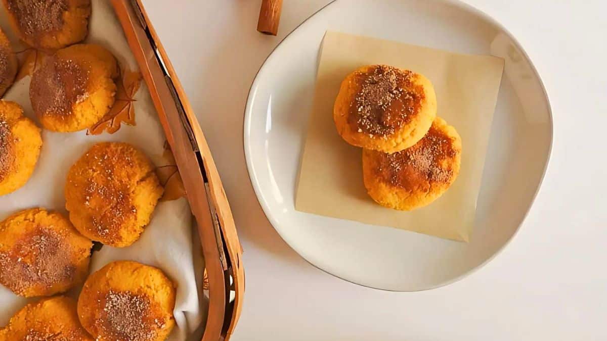 A white plate holds two cookies covered with cinnamon sugar beside a wooden basket filled with more cookies. The treats have a golden brown color and are placed on a light background.