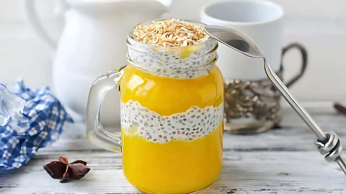 A mason jar filled with layers of creamy chia pudding and vibrant yellow mango puree, topped with toasted coconut flakes. A spoon rests on the jar's handle. The background features a white pitcher and mug on a light wooden surface.