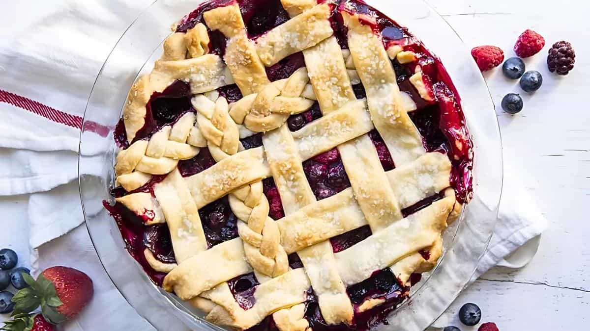 A freshly baked berry pie with a golden lattice crust. The pie is surrounded by scattered berries on a white surface, next to a white cloth with red stripes.