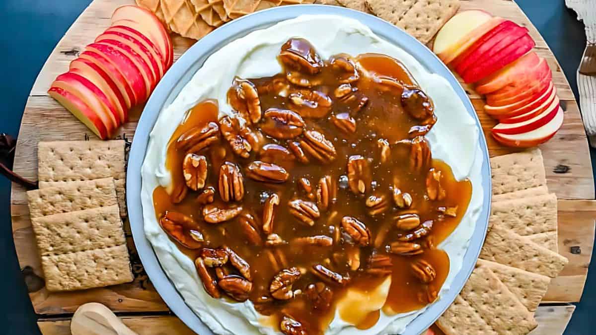 A round platter with cream cheese topped with caramel and pecans. Surrounding the platter are slices of red apples and golden crackers, neatly arranged on a wooden board.